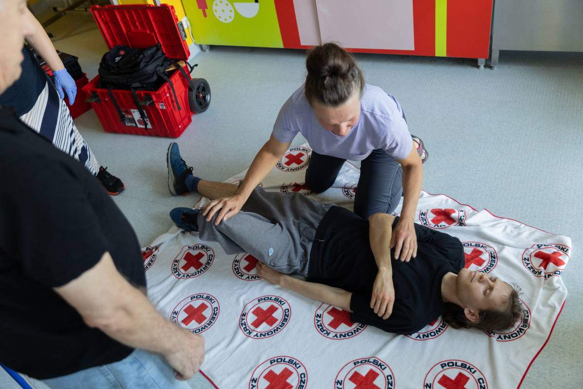 Kobieta pochylająca się nad mężczyzną, przy którym ćwiczy udzielanie pierwszej pomocy.